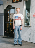 Man standing in front of a store wearing the Pieces No Vacancy GBP Rhinestone Tee Shirt Off White garment dyed with screen printed logo of the American flag, cut and sewn garment, boxy cropped fit, handset cross rhinestone pattern, hand distressed details Pieces GBP Rhinestone Tee Off White - Image 4 | Authenticated at Pieces Los Angeles