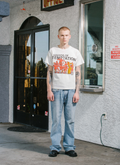 Man standing outside of a store facing the camera, wearing the Pieces Temptations Tee Shirt Off White with black text that says Sounds of Temptation with a graphic of red devils playing golden instruments in an orchestra band Pieces Temptations Tee Off White - Image 4 | Authenticated at Pieces Los Angeles