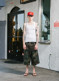 Man standing in front of store, wearing Pieces Back Pleat Shorts Dirty Camo distressed with a white tanktop and a red hat, facing the camera Pieces Back Pleat Shorts Dirty Camo - Image 6 | Authenticated at Pieces Los Angeles