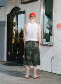 Man standing in front of store, wearing Pieces Back Pleat Shorts Dirty Camo distressed with a white tanktop and a red hat facing away from the camera Pieces Back Pleat Shorts Dirty Camo - Image 7 | Authenticated at Pieces Los Angeles