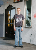 man standing in front of store facing the camera wearing the Pieces Future Tee shirt Vintage Black with blue jeans Pieces Future Tee Vintage Black - Image 5 | Authenticated at Pieces Los Angeles