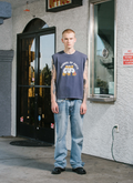 Man wearing Pieces Mascot Sleeveless Shirt Vintage Navy tank top with text that says School of Arts standing in front of a store facing the camera Pieces Mascot Sleeveless Shirt Vintage Navy - Image 4 | Authenticated at Pieces Los Angeles