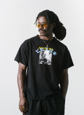 Man with a white background wearing the Pieces Post Card Tee shirt Black with a small faded white graphic of a city with yellow and light blue text over it, front view Pieces Post Card Tee Black - Image 3 | Authenticated at Pieces Los Angeles
